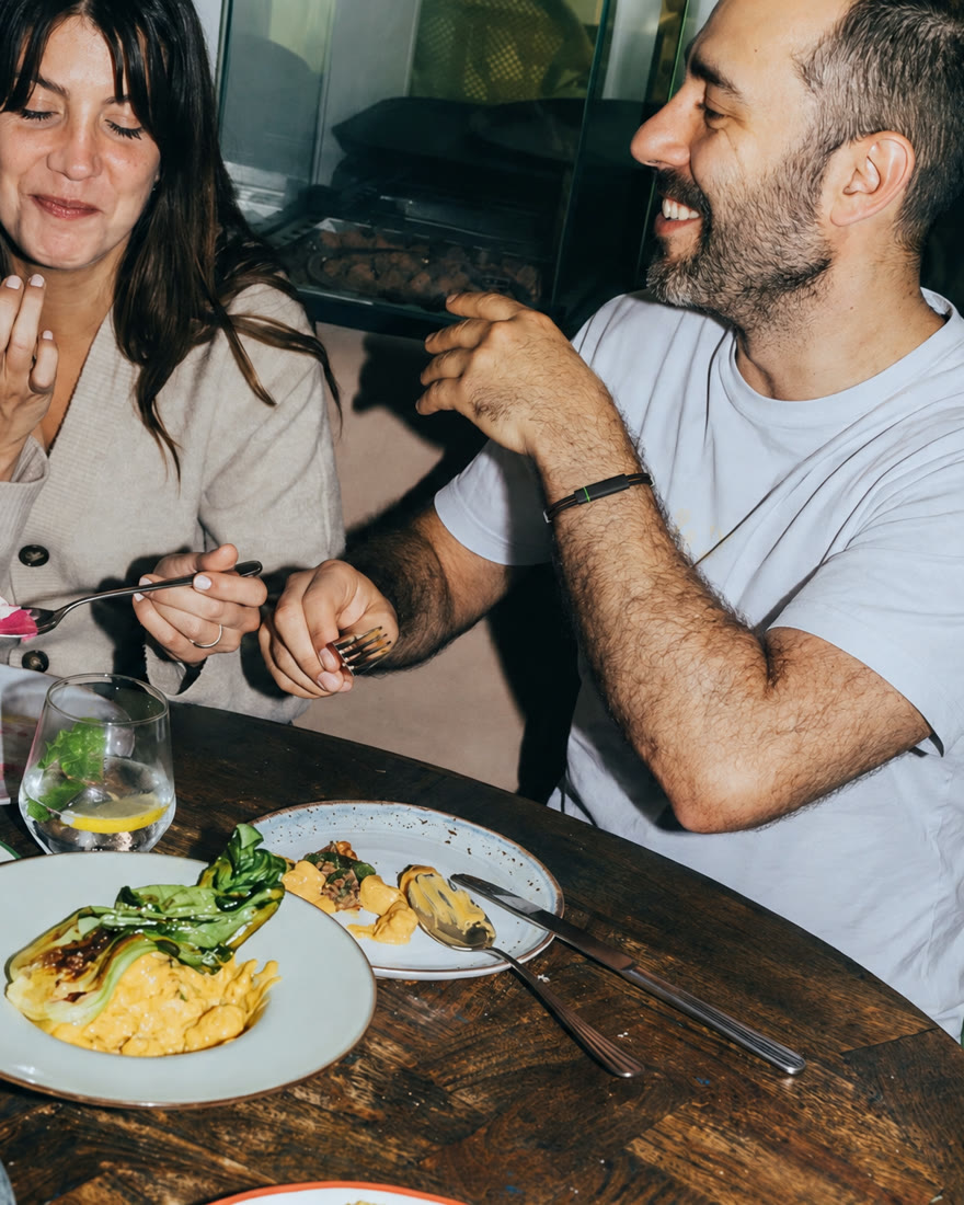 Two friends laughing over lunch at a wooden table full of shared plates